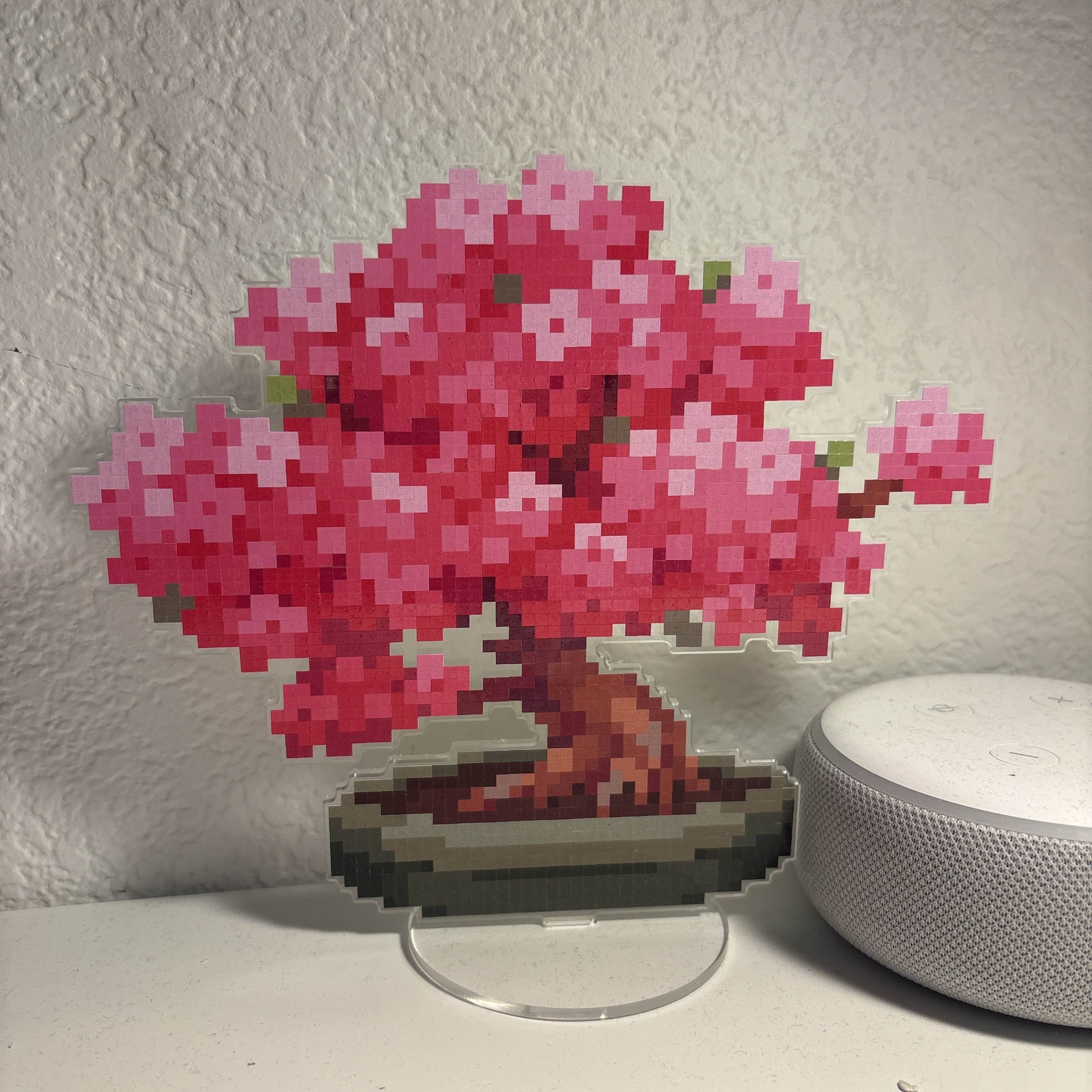 Pixelated cherry blossom tree on a stand next to a smart speaker on a white surface.